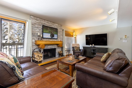 A cozy living room with leather sofas, a stone fireplace, a large TV, and a second TV on the wall. A window with drawn curtains shows a snowy outdoor scene.