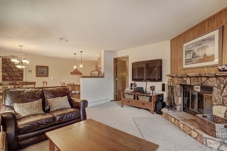 A cozy living room with a leather couch, wooden coffee table, stone fireplace, and wall-mounted TV. A dining area and kitchen with a breakfast bar are visible in the background.