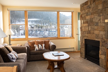 A cozy living room with a stone fireplace, large windows showing a snowy mountain view, and a brown sofa with pillows. A round wooden coffee table holds a closed laptop and a decorative object.