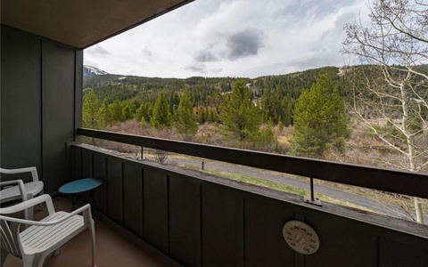 A balcony with two white chairs and a small blue table overlooks a scenic view of a forested mountainside.