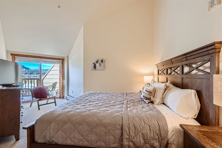 A cozy bedroom with a large bed, wooden headboard, and bedside tables with lamps. There is a chair, a dresser, and a balcony with a mountain view in the background.