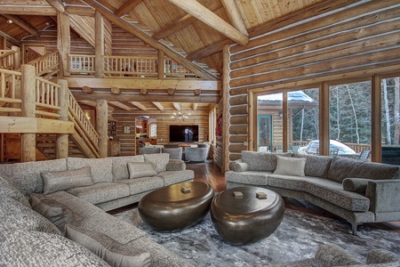 A spacious, modern log cabin living room with large windows, beige sofas, round metallic coffee tables, and a loft above. The room features exposed wooden beams and overlooks a snowy outdoor scene.