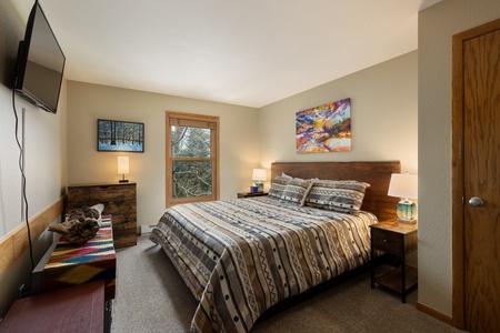 A bedroom with a large bed featuring patterned bedding, two bedside tables with lamps, a flat-screen TV mounted on the wall, and framed artwork on the beige walls. A window lets in natural light.