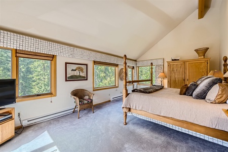 A spacious bedroom with large windows that offer a view of trees, a wooden bed with beige bedding, a wicker chair, a TV, and wooden furniture including a dresser and side tables.