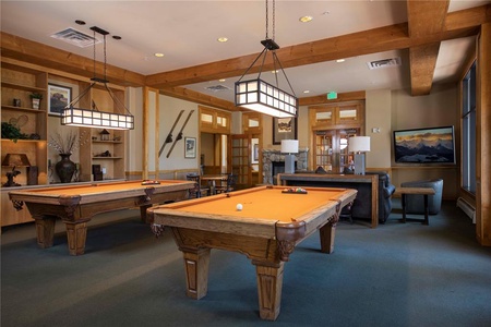 A recreational room with two wooden pool tables, hanging lights, a seating area with a sofa and armchairs, and wall decor, including skis and framed artworks.