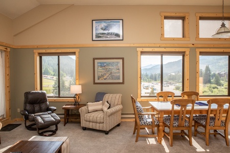 A living room with a recliner, an armchair, a side table, and a dining table with six chairs. Large windows offer a view of a mountainous landscape. Two framed pictures hang on the wall.