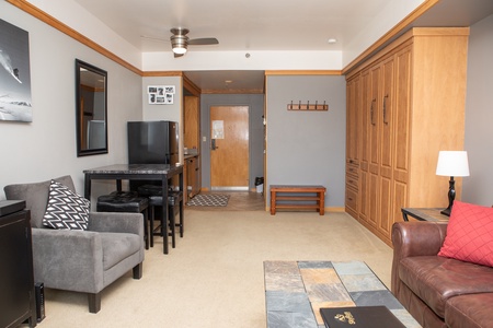 A small living area with a gray armchair, a brown leather chair, a table with stools, a mini-fridge, and wooden cabinetry. The floor is carpeted, and there is a door at the far end of the room.