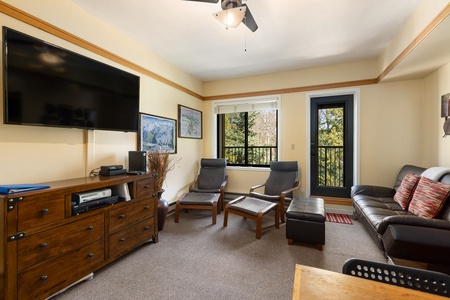 Living room with a mounted flat-screen TV, two lounge chairs with footrests, a black leather sofa, a wooden chest of drawers, and a balcony with a view of trees.