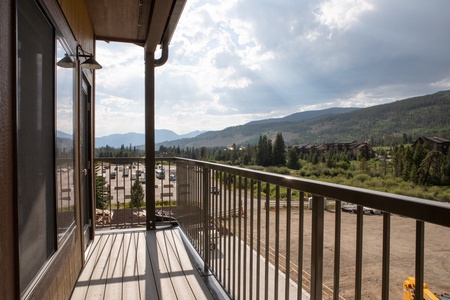 A balcony with metal railing overlooks a parking area and distant forested mountains under a partly cloudy sky.