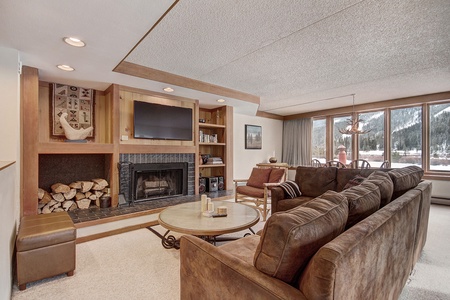 Cozy living room with a fireplace, bookshelves, a brown couch, coffee table, and large windows with a mountain view. Adjacent dining area with a table and chairs. Wood logs stored by the fireplace.