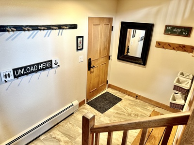 Stairwell landing with a wooden door, a large mirror, coat hooks, and a "UNLOAD HERE" sign. Baskets on the right hold items. Energetic decor includes "EAT, SLEEP, SKI" sign.