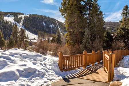 A wooden footbridge over snow leads to a mountain with ski trails and pine trees under a clear blue sky.