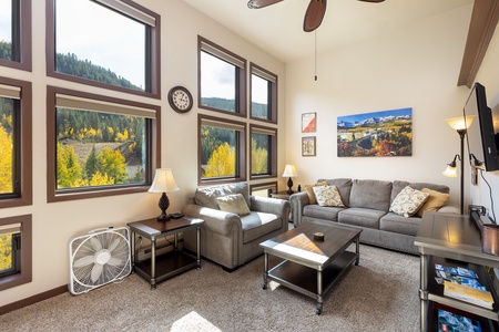 A living room with large windows, grey sofas, a coffee table, two side tables with lamps, a ceiling fan, a wall clock, a fan, and autumn landscape decor. Trees and hills visible outside.