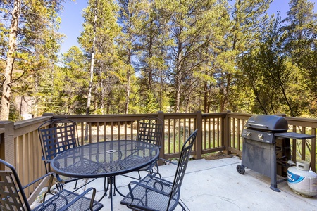 Outdoor deck with a black metal table and four chairs set, a gas grill, and surrounded by tall pine trees.