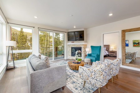 A modern living room with large windows, a grey sofa, patterned armchairs, a TV above the fireplace, and a blue chair. The room features a view of trees and mountains outside.