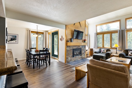 A well-lit living and dining area with wooden flooring, a fireplace, a wall-mounted TV, a dining table with six chairs, and large windows. There is a door to an outdoor area beneath a hanging light fixture.