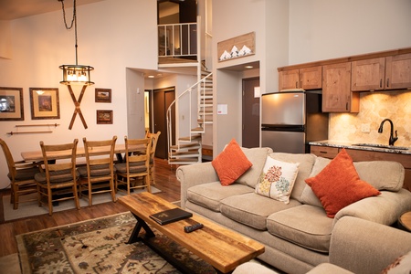 A cozy living area featuring a beige sofa with orange pillows, a wooden coffee table, and a nearby dining table with chairs. The kitchen has wooden cabinets and a spiral staircase leads to the upper level.