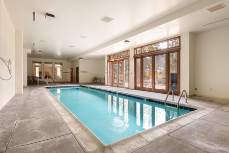 Indoor swimming pool with concrete decking and lounge chairs along one side. Large windows and glass doors on one wall provide natural light and a view of an outdoor area.