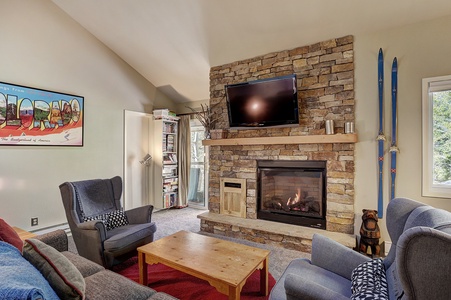 A cozy living room features a stone fireplace with a TV on top, a wooden coffee table, plush seating, a colorful wall poster, a shelf with books, and skis leaned against the wall. Light fills the room.