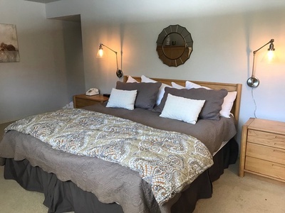 A neatly made bed with gray and white pillows, a patterned bedspread, two bedside tables with wall-mounted lamps, and a round mirror above the headboard in a well-lit bedroom.