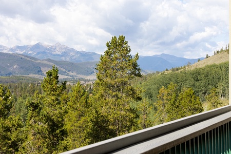 View of forested mountains with a cloudy sky, seen from a balcony with a wooden railing in the foreground.