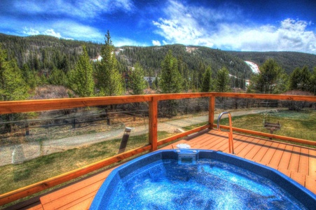 A blue hot tub is situated on a wooden deck overlooking a scenic mountainous landscape with pine trees and a partly cloudy sky.