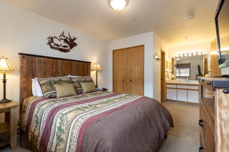 A cozy bedroom with a wooden bed frame, patterned bedding, two bedside lamps, a dresser, a wall-mounted TV, and an ensuite bathroom. A wall decoration of a deer head silhouette is above the bed.