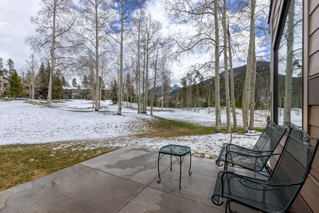 A snow-dusted patio with two metal benches and a small table overlooks a wintry landscape of sparse trees and distant mountains under a partly cloudy sky.