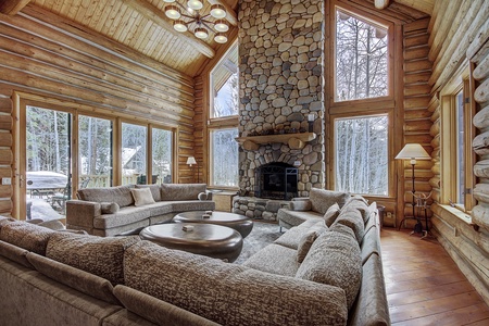 Spacious living room with large windows, a stone fireplace, and a wrap-around gray sectional sofa in a log cabin. Snow-covered trees are visible outside.