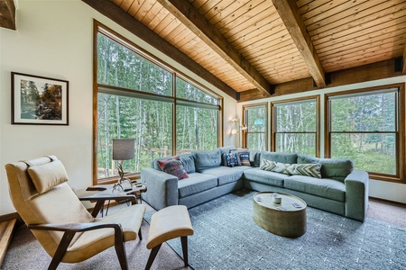 A cozy living room with a large corner sofa, reclining chair, round coffee table, and large windows offering a view of lush greenery. The room has a wooden ceiling with exposed beams.