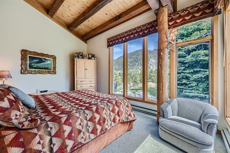 A cozy bedroom with a large bed, wooden furniture, a patterned bedspread, an armchair, and a large window offering a scenic view of mountains and trees.