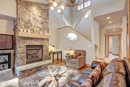 A cozy living room with a stone fireplace, brown leather sofa, armchair, glass coffee table, rug, wooden ceiling fan, and staircase leading to an upper level.