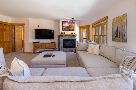 A living room with a large sectional sofa, a TV on a wooden stand, a fireplace with a painting above it, and multiple wooden doors and windows.