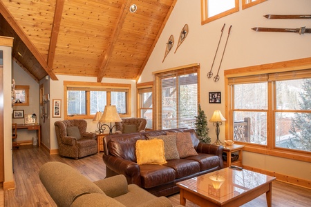 A cozy living room with a wooden ceiling, brown leather sofa, armchairs, coffee table, lamps, and wall decorations including old skis and snowshoes. Large windows offer a view of snowy trees outside.