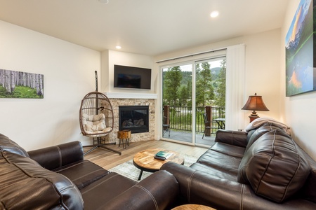 A cozy living room with a sectional leather sofa, a round coffee table, a hanging chair, a fireplace with a TV above it, and a large sliding glass door leading to an outdoor patio with a forest view.