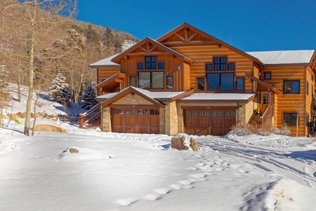 A large, wooden cabin-style house with multiple garages is surrounded by snow, trees, and hills under a clear blue sky.