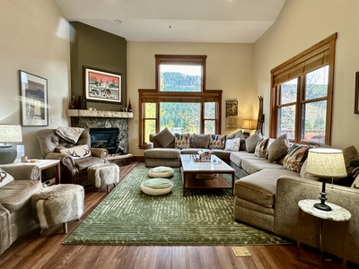 A cozy living room with a large sofa, green rug, and fireplace. Mountain views are visible through the windows.