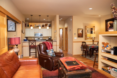 A cozy living room with a tan leather couch and recliner, an open kitchen with pendant lights, a dining area, and wooden flooring throughout. The room features warm lighting and decorative accents.