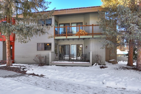 Two-story house with a balcony and sliding glass door, surrounded by snow-covered ground and pine trees. Firewood is neatly stacked on the balcony. The house has a light gray exterior.