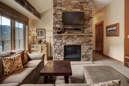 A cozy living room with a stone fireplace, mounted TV, sectional sofa, wooden coffee table, and a window with a view of the mountains.