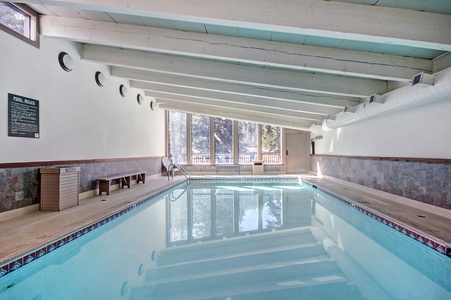 Indoor swimming pool with blue water, surrounded by white walls and large windows letting in natural light. Ceiling has exposed beams and lights. A few chairs are placed along one side.