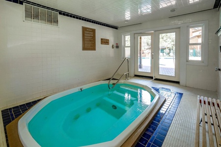 Indoor hot tub with blue tiles is surrounded by white walls and ceiling, adjacent to glass doors leading to an outdoor area. A bench is placed beside the hot tub. Instructions are posted on the wall.