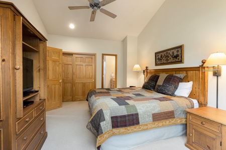 A bedroom with a patchwork quilt on a double bed, wooden furniture, a ceiling fan, and a nightstand with a lamp. The room has an open door leading to another area.