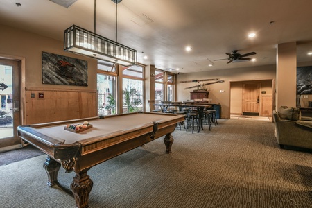 A recreation room with a pool table, seating area, and wooden décor. Large windows on one side and a ceiling fan are visible. Bright overhead lighting illuminates the space.