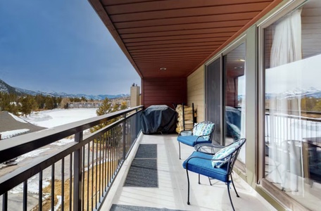 A covered balcony with two blue chairs and a barbecue grill, looking out over a snowy landscape with mountains in the distance.