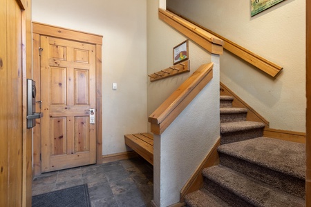 A wooden front door and carpeted staircase in a home. The foyer includes a small bench and a wall-mounted coat rack. A framed picture hangs above the bench.