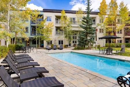 Outdoor pool area with lounge chairs, tables, and umbrellas, surrounded by trees and multi-story residential buildings.