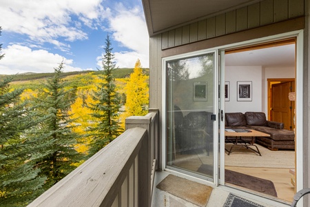 View of a cozy living room through an open sliding glass door, with a balcony railing in the foreground and trees with yellow and green foliage in the background.