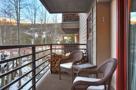 A balcony with two wicker chairs and a small table overlooks a snowy wooded landscape, with firewood neatly stacked against the wall.