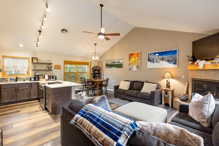 Open-concept living room with a kitchen area, dining table, couches, fireplace, and large windows. Decor includes framed landscape photos and a ceiling fan.